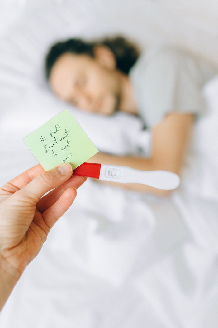 A woman's hand holding a positive pregnancy test and a note with a person sleeping in the background.